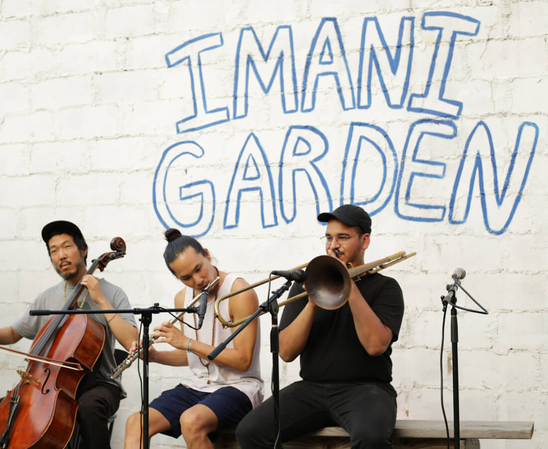 Three musicians playing instruments in front of a wall at Imani Community Garden