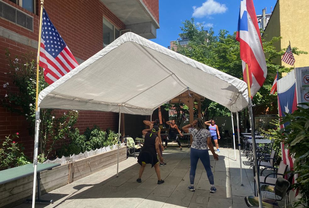 A group of people participating in a zumba class with instructor Frances under an awning with an American flag and a Puerto Rican flag